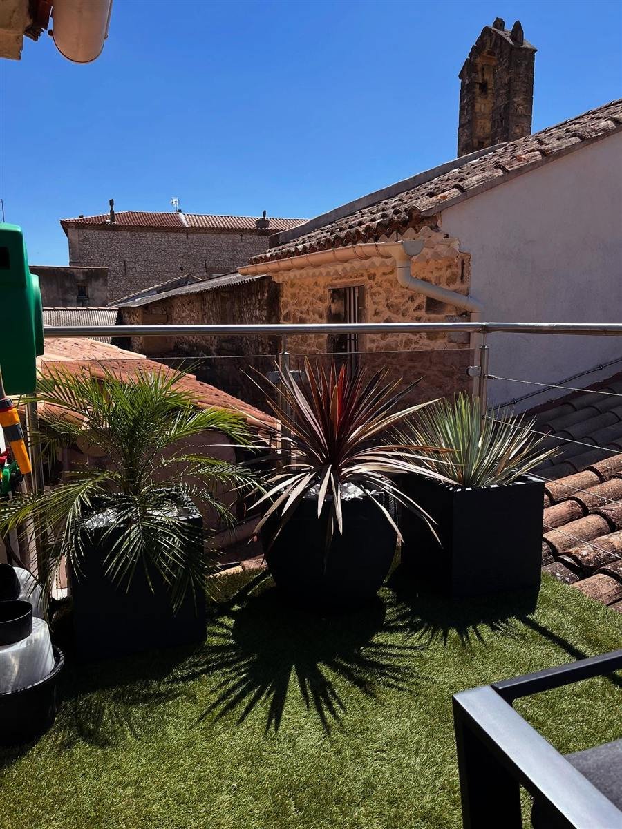 Aménagement d’un toit-terrasse par UnionVerte à Villeneuve-lès-Avignon : palmier, cordyline pourpre et yucca en bacs noirs sur gazon synthétique, face aux toits de tuiles du village.